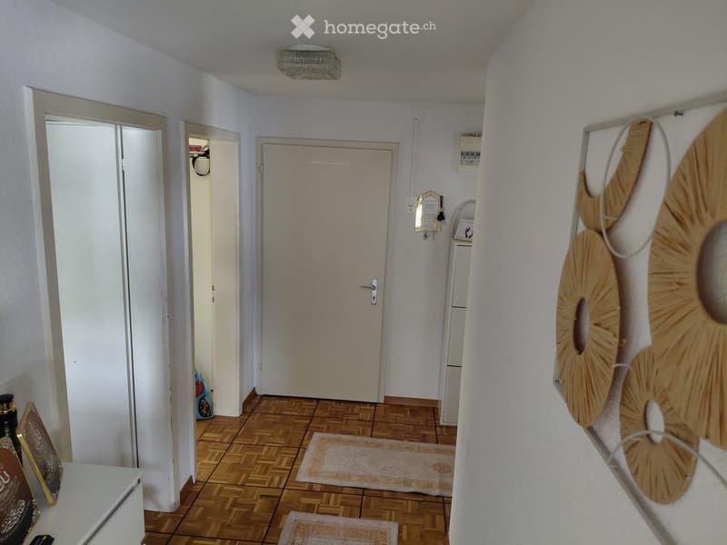 A narrow hallway with wooden flooring, white walls, and several doors leading to different rooms. A large decorative wall piece is visible on the right side.