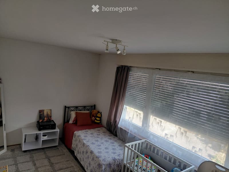 A cozy bedroom with a bed, nightstand, mirror, and a small cradle. The room features large windows with blinds and curtains, and a picture frame on the nightstand.