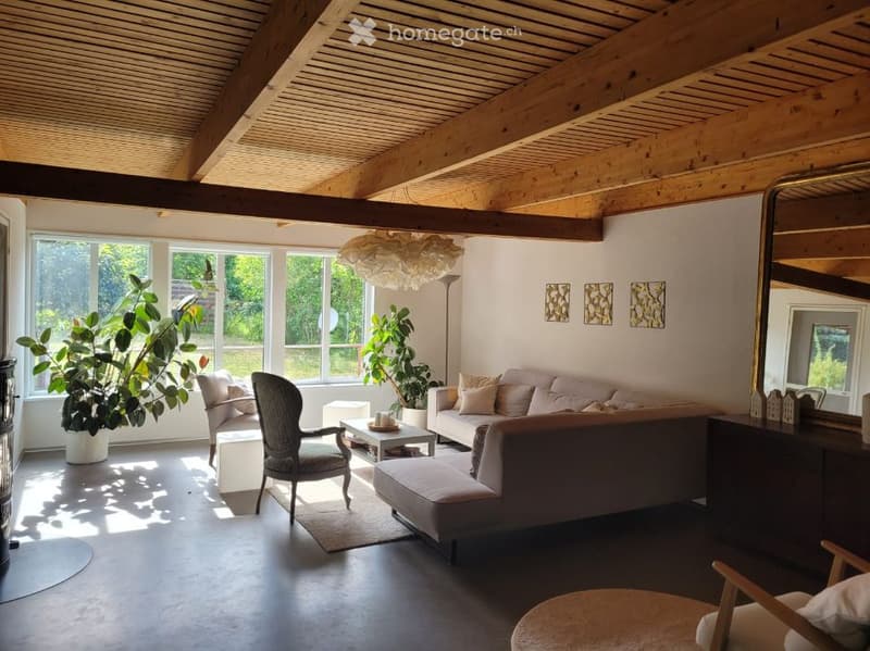 Bright living room with large windows, exposed wooden beams, a sofa, armchair, coffee table, ottoman, floor lamp, and decorative mirror.