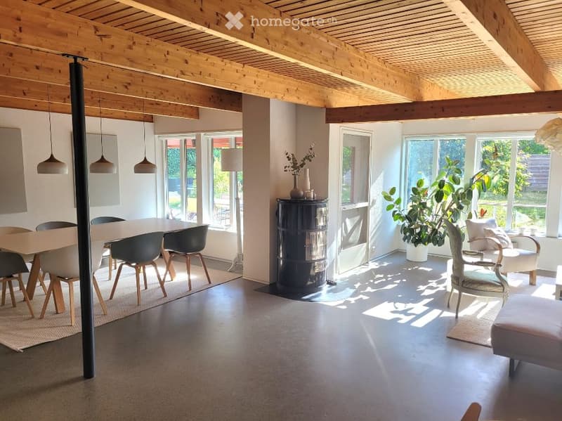 Open-plan living and dining area with wooden ceiling beams, large windows, modern furniture including a dining table, chairs, sofa, ottoman, and a floor lamp.