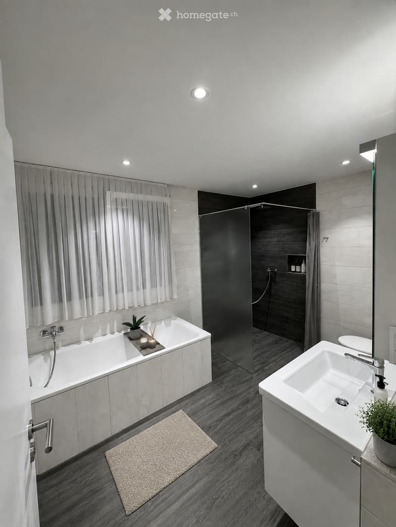 Modern bathroom with a white bathtub, double sink, large mirror, and a separate shower area with dark tiles. Features include a small plant, candles, and a beige rug on the floor.
