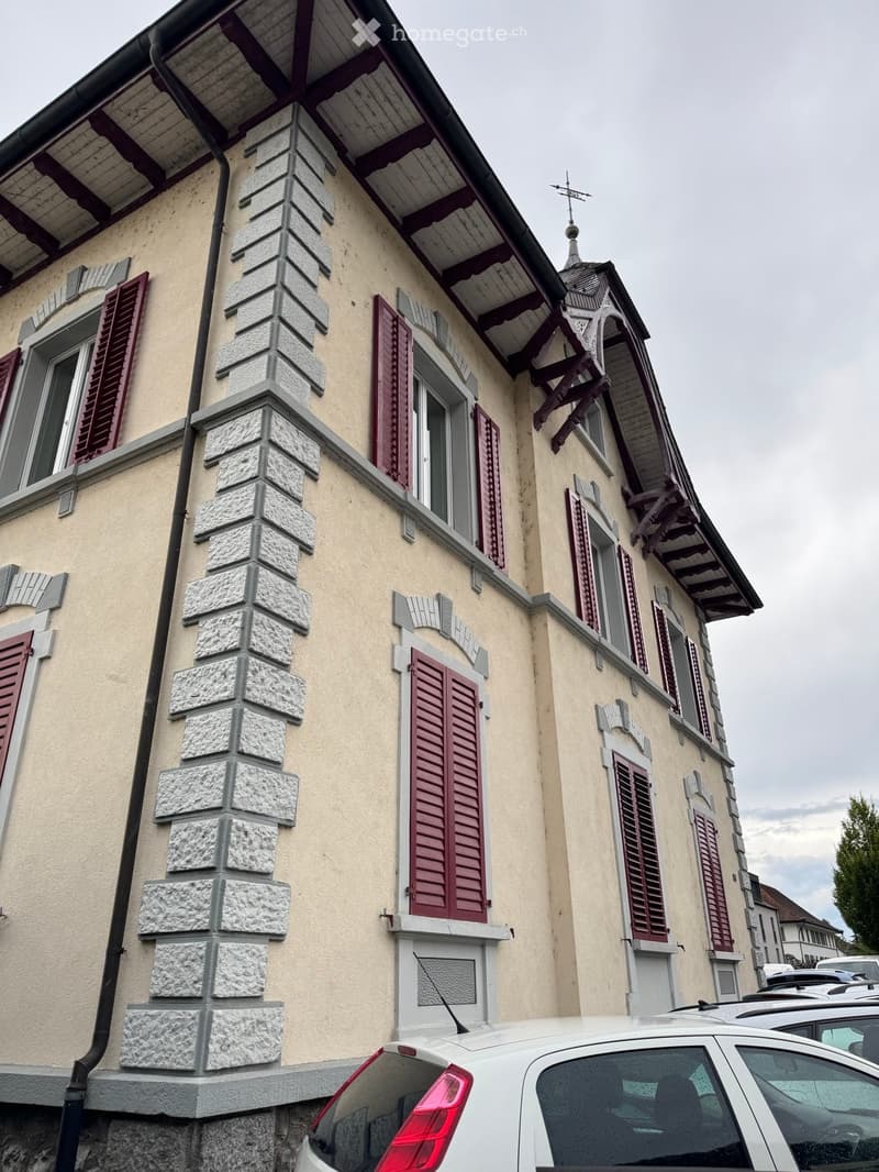 Two-story house with beige walls, stone accents, red shutters, and a decorative roofline.