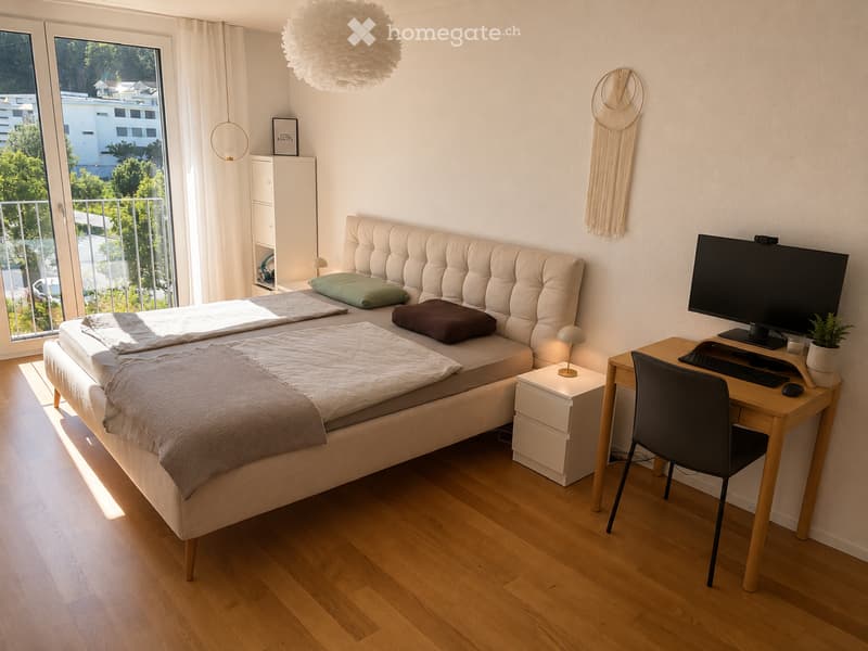 Bright bedroom with a large bed, a desk with a computer, a nightstand, and a view of a balcony. The room features wooden flooring, white walls, and decorative elements like a dreamcatcher and a hanging light fixture.