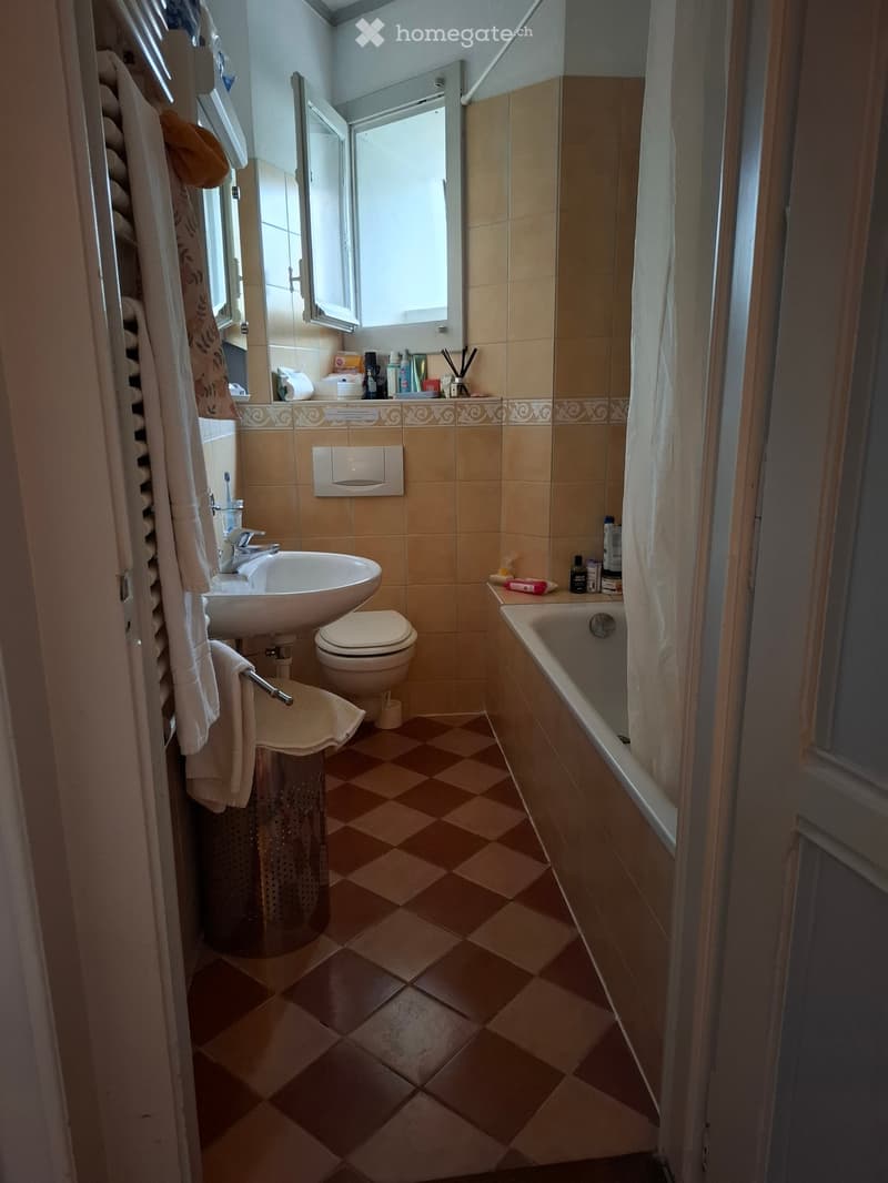 Bathroom with beige tiles, a sink, toilet, bathtub, towel rack, and a mirror. The room features a checkered floor and a window above the bathtub.