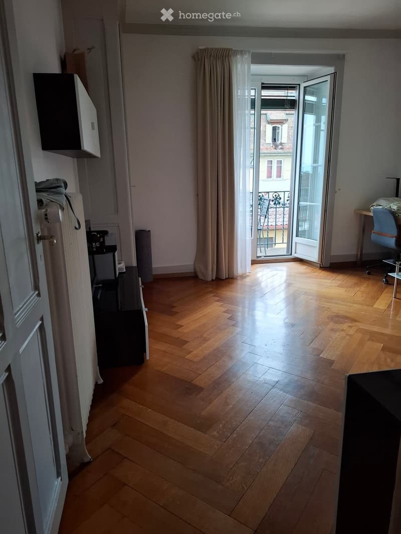 Empty room with wooden flooring, open sliding door leading to a balcony, and light-colored walls.