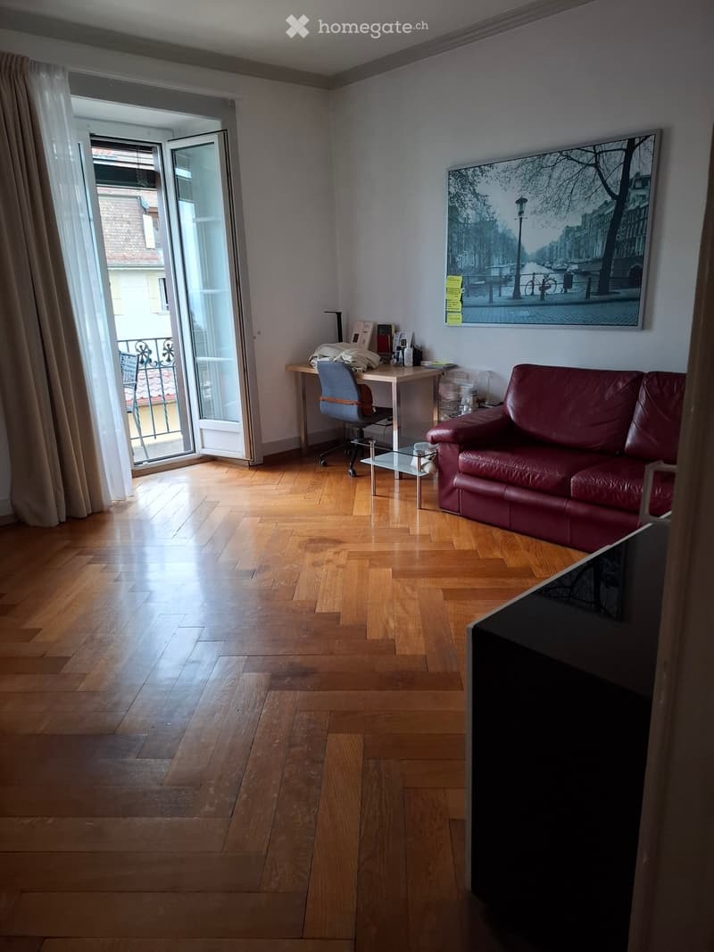 A furnished room with wooden flooring, a red leather sofa, a glass coffee table, a desk with a chair, and a large screen on the wall.