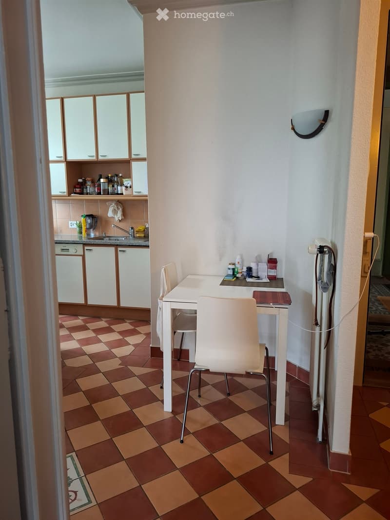 A small, well-lit room with a white table and chair, a refrigerator, a sink with a faucet, and several bottles on the counter. The floor is tiled in a checkered pattern of brown and beige.
