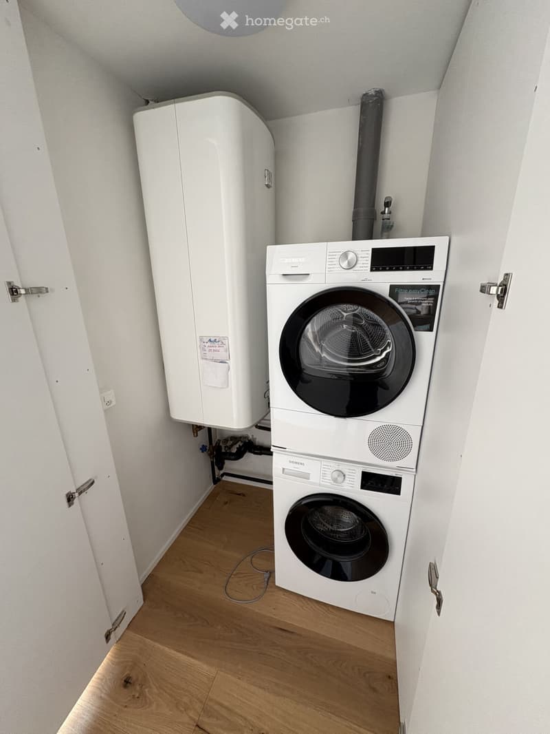 A small room with a white washing machine and a white dryer stacked on top of each other, a white water heater on the left, and a white door on the right.