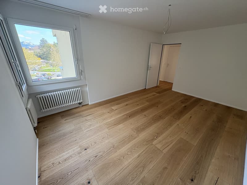 Empty room with wooden floor, white walls, large window, radiators, two doors