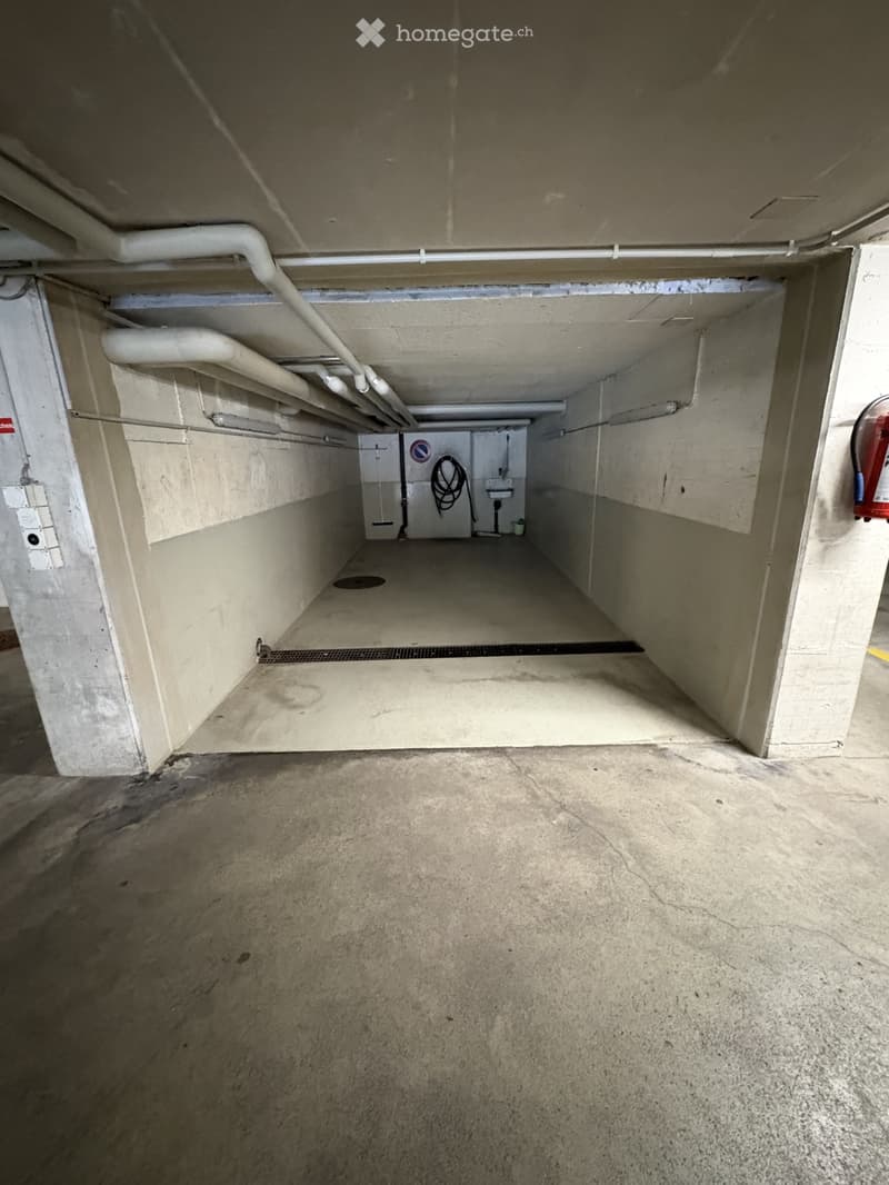 Empty parking space with white walls, concrete floor, pipes on the ceiling, and a fire extinguisher on the right wall.