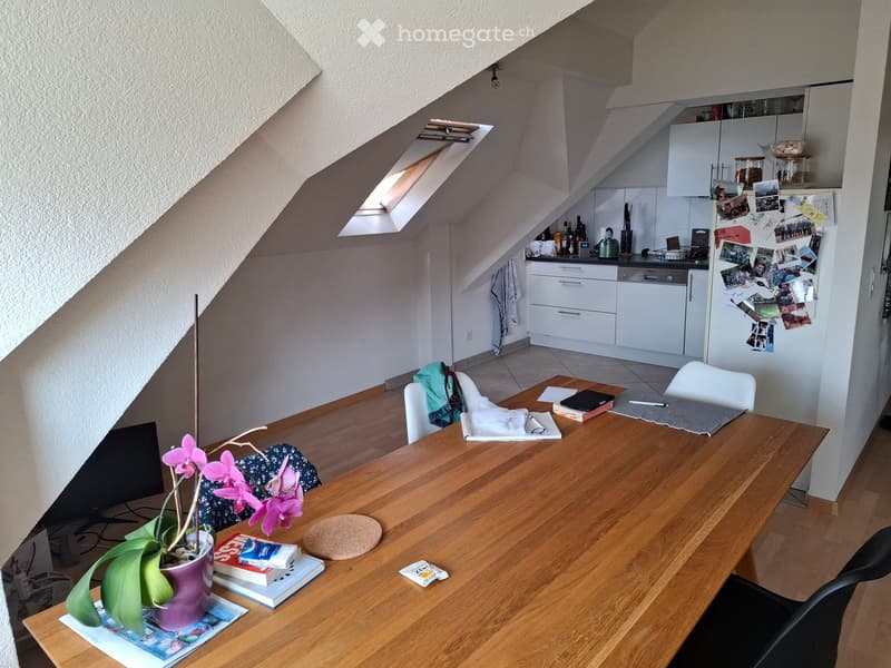 A cozy attic room with a wooden dining table, chairs, laptop, and a small kitchen area with white cabinets, a refrigerator, and a dishwasher. The room features a skylight and a slanted ceiling.