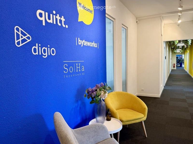 Modern hallway with a blue wall featuring company logos, a small round table with a vase of flowers and a bottle, and two chairs.