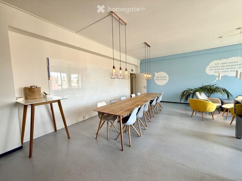 Modern, well-lit room with a long wooden table surrounded by chairs, a sofa, and a small plant. The space features pendant lights and a whiteboard on one wall.