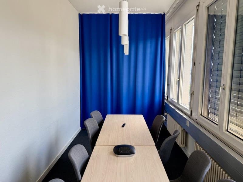 A modern, unfurnished room with a long wooden table surrounded by grey chairs, a blue curtain, and a hanging light fixture.