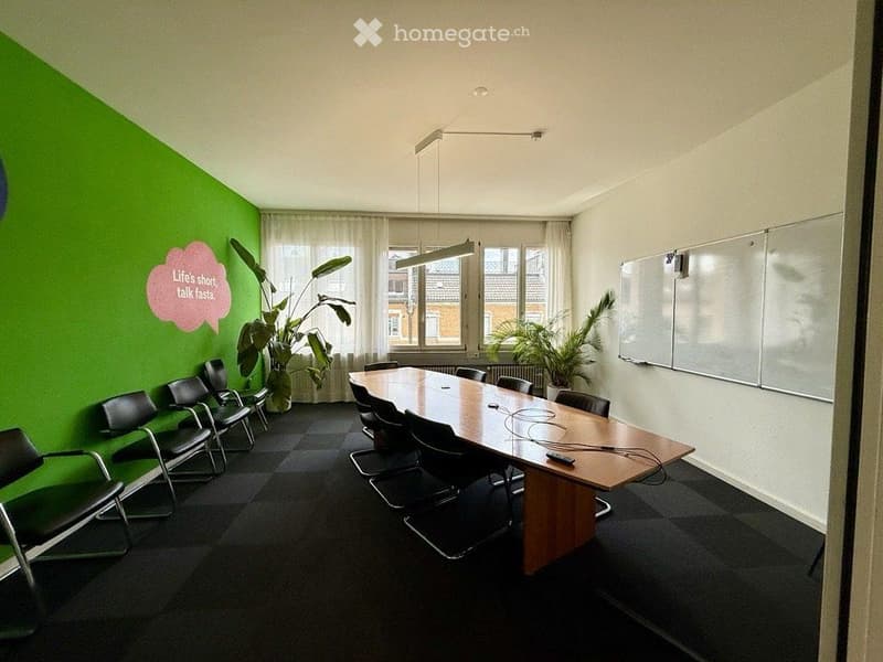 Bright conference room with a long wooden table surrounded by black chairs, two large potted plants, and a whiteboard on the wall.