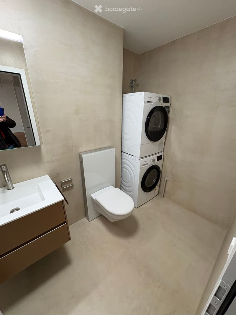 Laundry area with sink and mirror, modern bathroom fixtures, built-in white cabinetry, white tile flooring, two stacked Siemens washing machines, wall-mounted soap dispenser, toilet adjacent to washing machines, white ceramic toilet bowl.