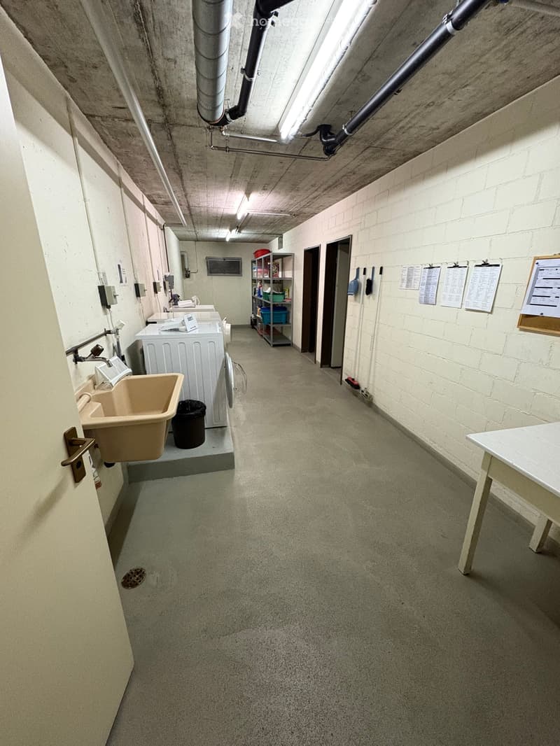 Empty laundry room with several washing machines, sink, garbage bin, white walls, shelves, and bulletin boards.