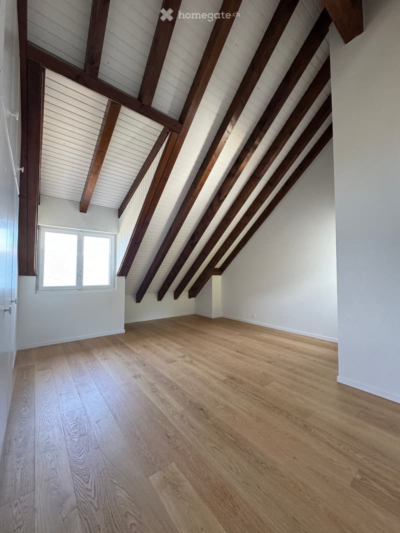 Empty attic space with exposed wooden beams, light wooden flooring, and a small window letting in natural light.