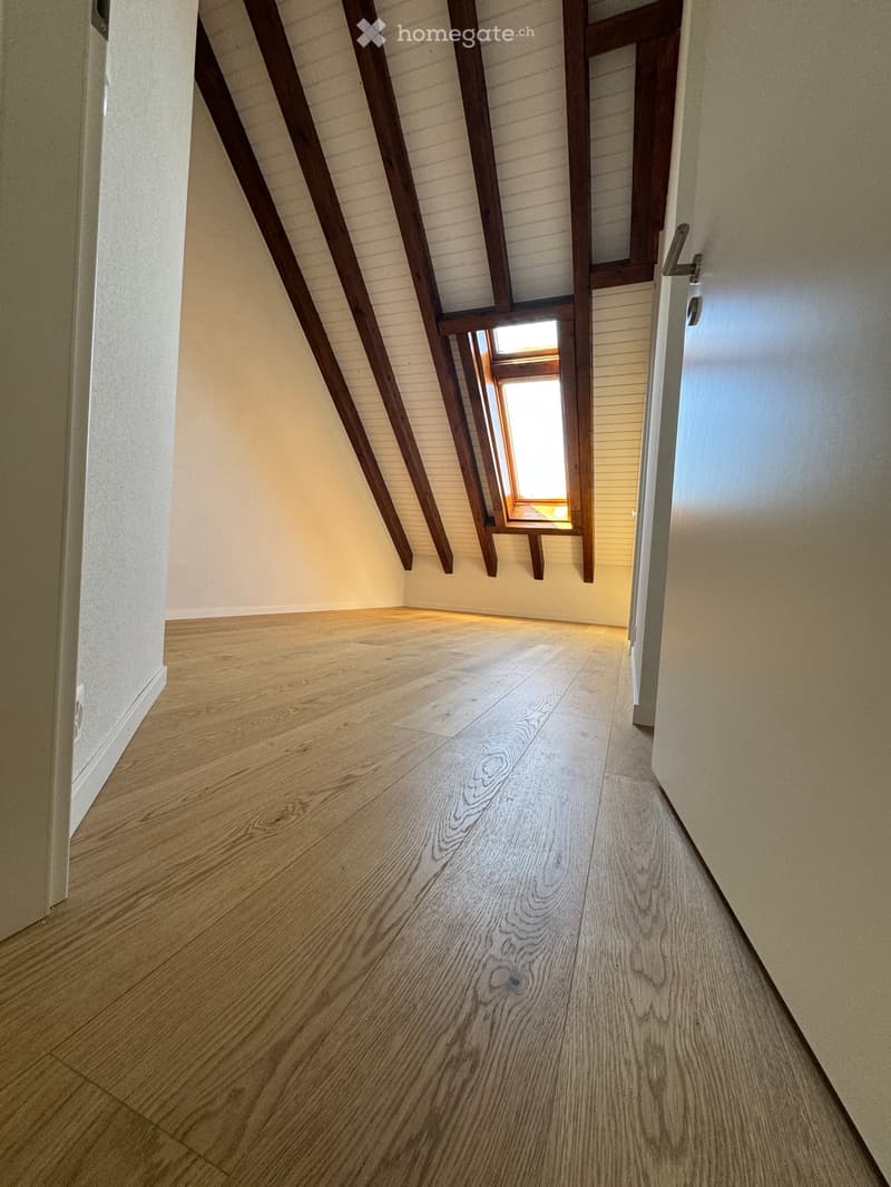 Empty hallway with wooden flooring, white walls, and exposed wooden beams on the ceiling. A small window allows natural light to enter.