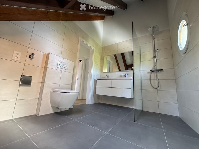 Modern bathroom with a toilet, sink, mirror, and glass-enclosed shower. Light-colored tiles and a dark grey floor.
