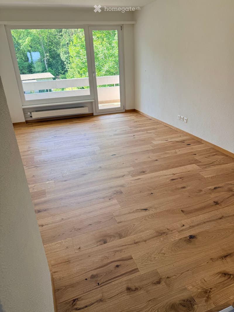 Empty room with light wooden flooring, white walls, large window and sliding glass door leading to a balcony.