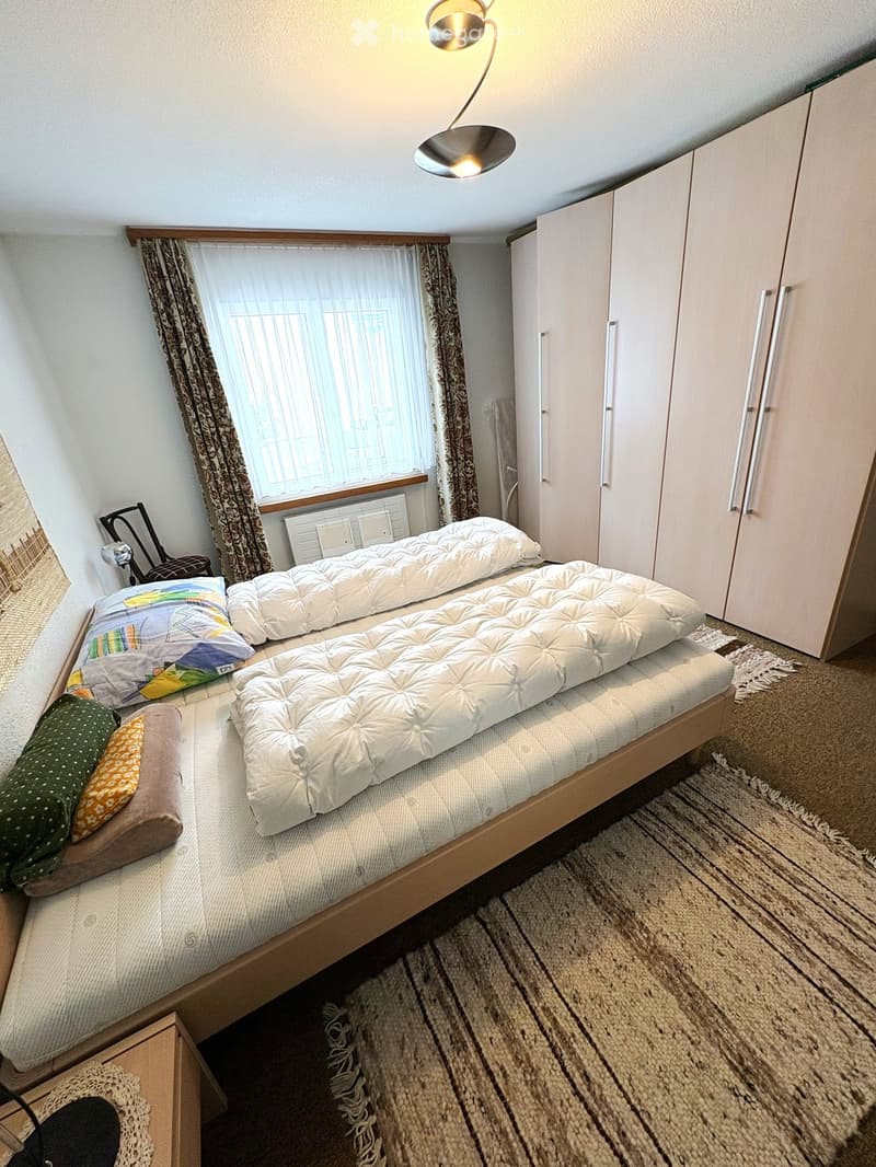 Cozy bedroom with a wooden bed, white quilted mattress, colorful pillows, a wardrobe, a chair, and a lamp.