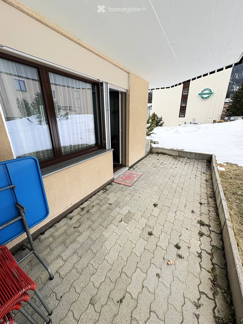 Balcony with tiled floor, two chairs (one blue, one red), window with curtains, and a small mat near the entrance.