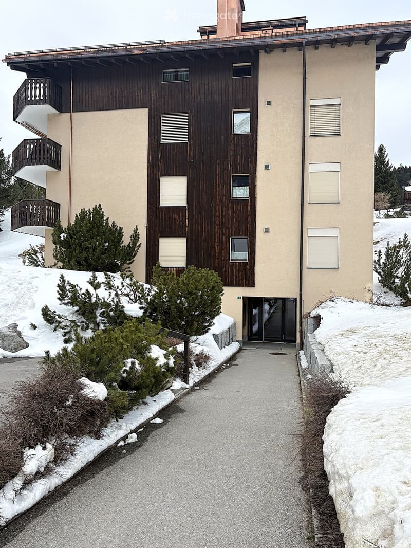 Two-story building with a wooden facade and beige walls, featuring multiple windows and balconies, surrounded by snow-covered ground and small evergreen trees.