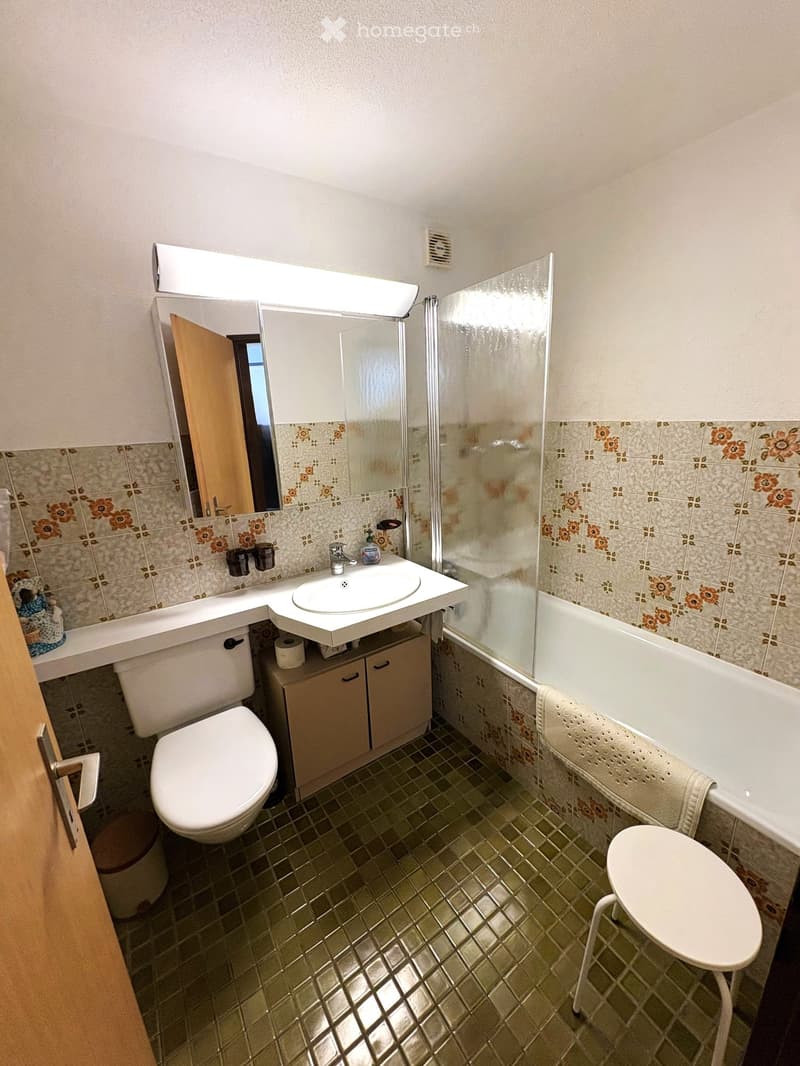 Bathroom with floral tiled walls, a white toilet, a sink with a mirror, a bathtub with a glass shower enclosure, and a small stool.