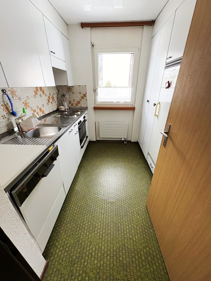 Compact kitchen with white cabinets, stainless steel sink, oven, and a window with a curtain. Green hexagonal floor tiles.