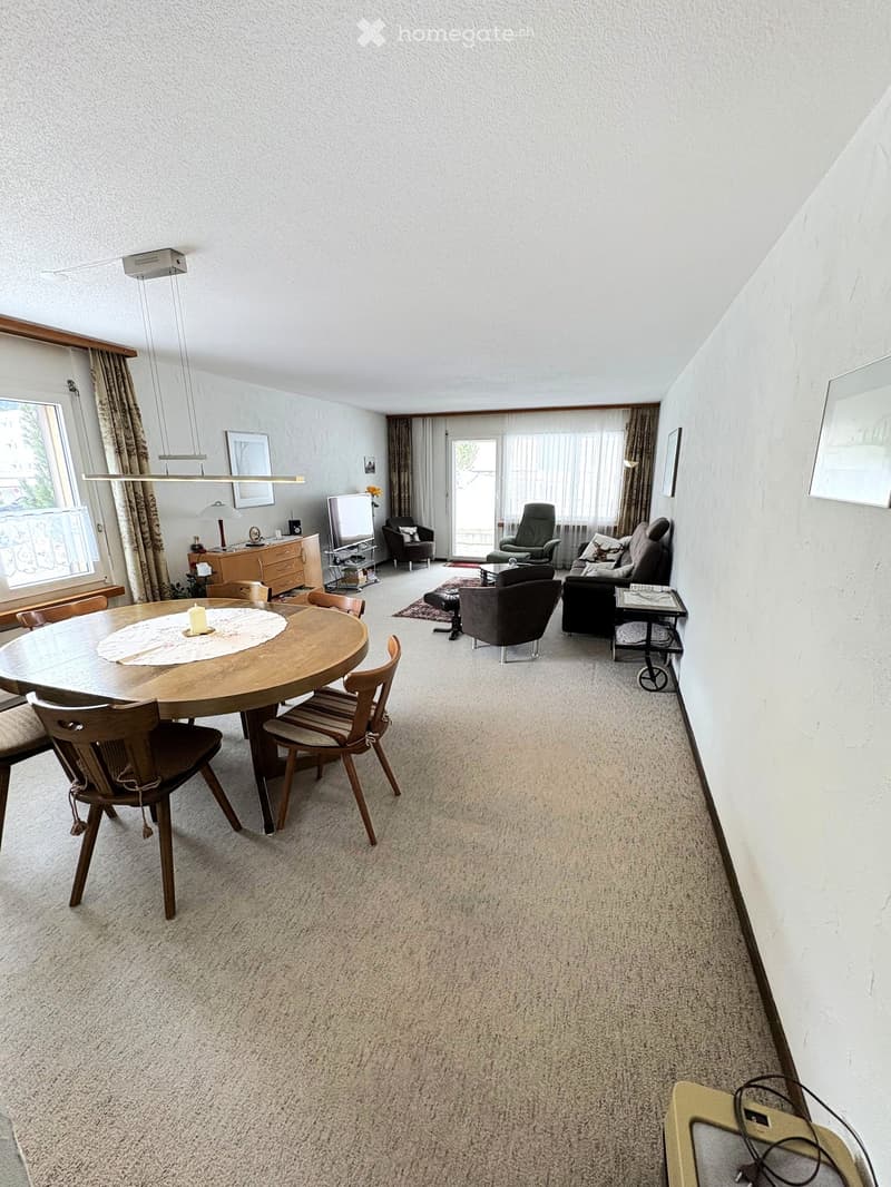 A well-lit living and dining area with a round wooden dining table, chairs, a sofa, armchairs, a television, and a cabinet with a lamp.