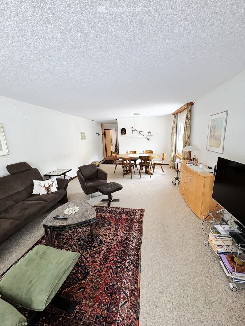 A spacious living room with a brown sofa, a glass coffee table, a green ottoman, a TV cabinet, and a dining area with a round table and chairs.