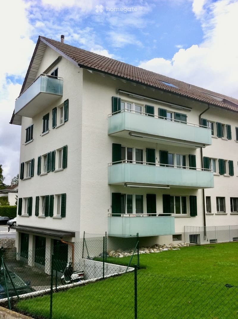 Modern three-story building with white facade, multiple balconies, green shutters, and a fenced lawn area.