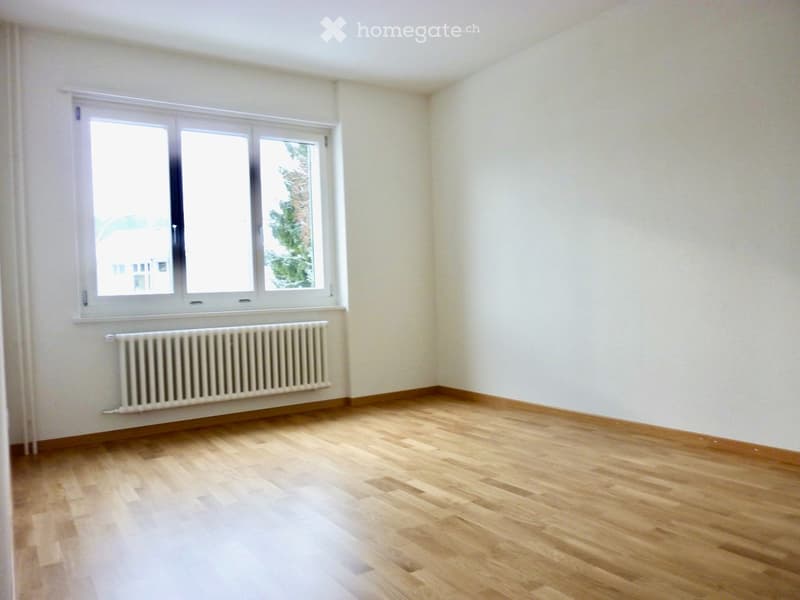 Empty room with wooden flooring, white walls, and a large window with white frame. A radiator is visible below the window.