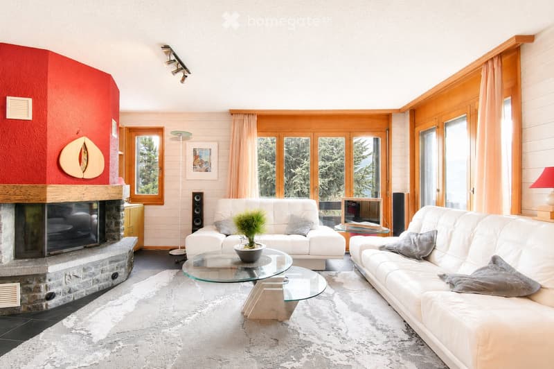 Modern living room with white sofas, a glass coffee table, a stone fireplace, and large windows with wooden frames. The space features a white shag rug and a potted plant on the coffee table.