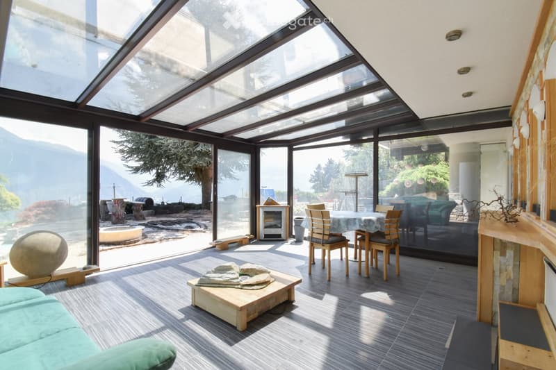 Bright, airy room with large glass walls and a skylight, featuring a green sofa, wooden coffee table, dining area with chairs and table, and a small fireplace. The space has a modern, minimalist design with natural light and a scenic mountain view.