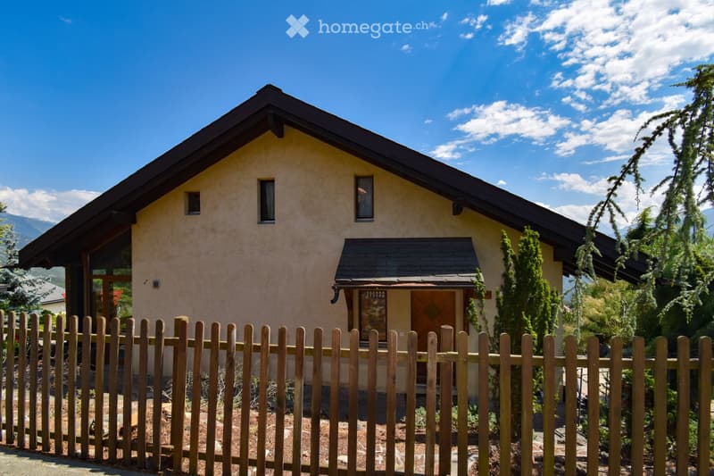 Single-story house with beige stucco walls, dark roof, and wooden front door. Surrounded by a wooden fence and greenery, with mountains visible in the background.