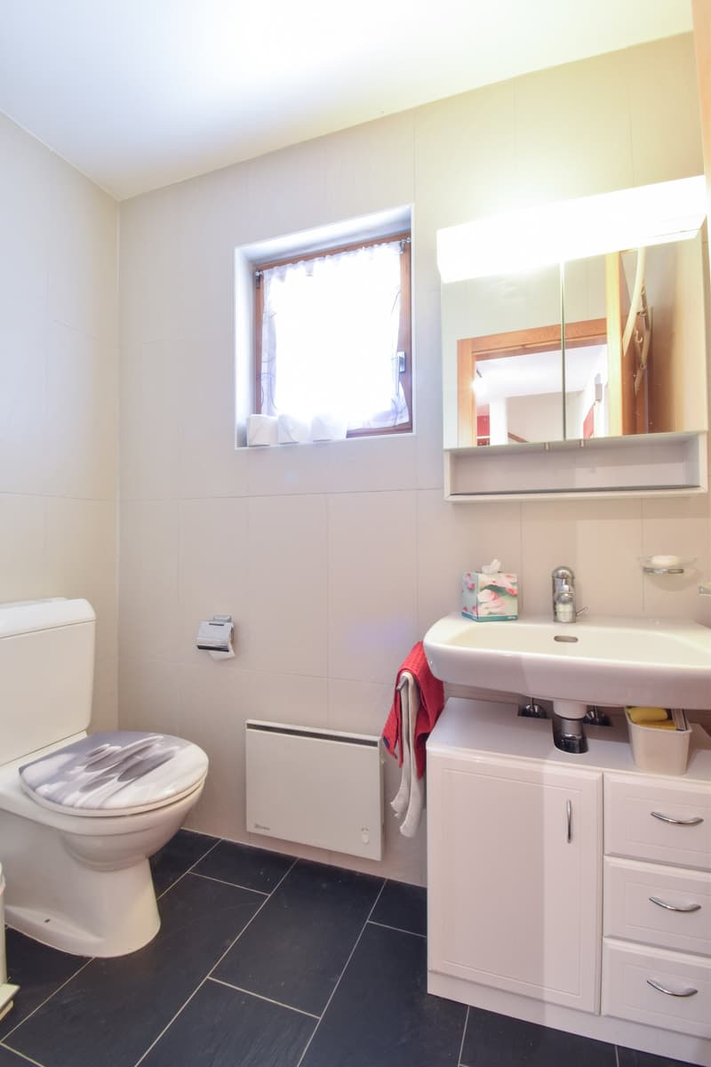 Modern bathroom with white tiles, black floor, toilet, sink with cabinet, mirror, towel, radiator, and window with curtain.