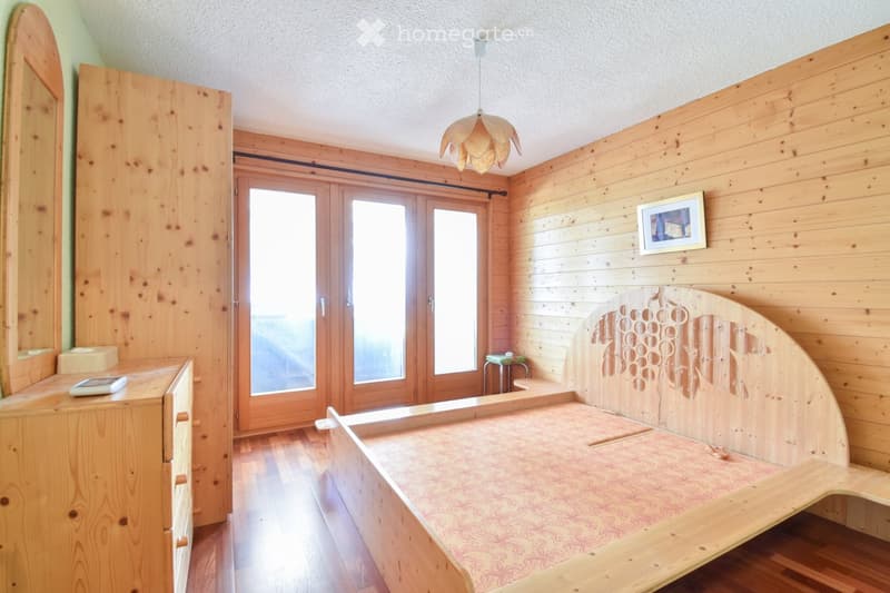 Cozy wooden bedroom with a large bed, nightstand, mirror, and a small stool near the window. The room features wooden walls and a hanging light fixture.