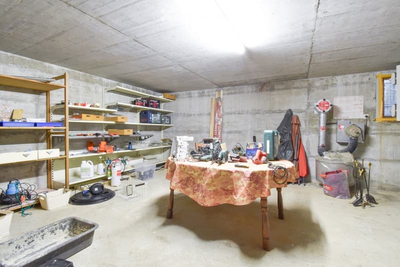 A basement or utility room with concrete walls and ceiling, containing a wooden table with a patterned tablecloth, shelves filled with tools and supplies, and various storage items.