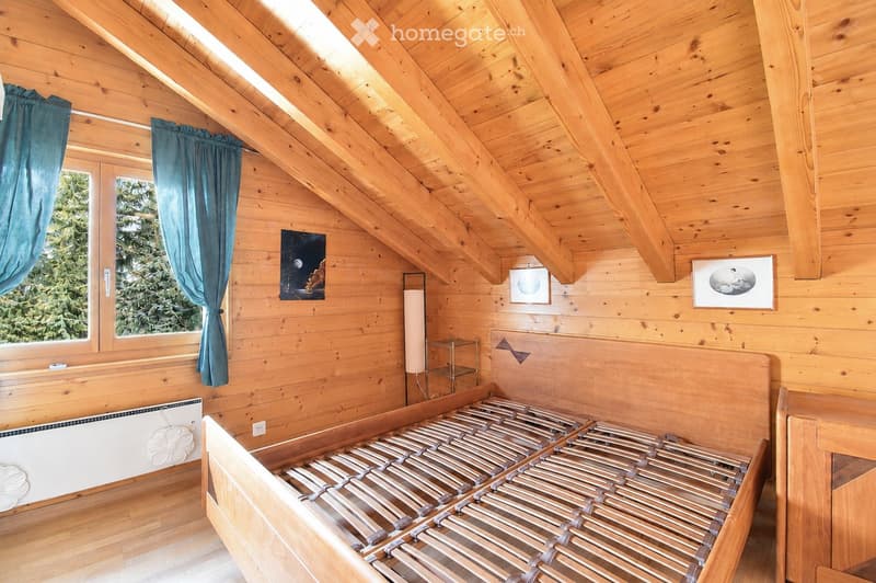 Cozy wooden bedroom with slanted ceilings, large window with blue curtains, two single beds, nightstand with lamp, and artwork on walls.