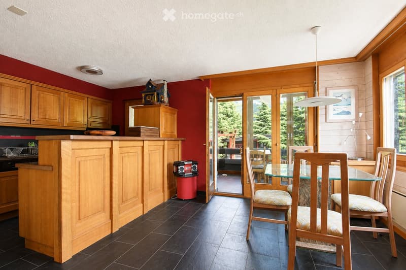 A modern kitchen with wooden cabinets, a central island, and a dining area with chairs and a glass table. The space features dark tiled flooring and large windows with a view of greenery.