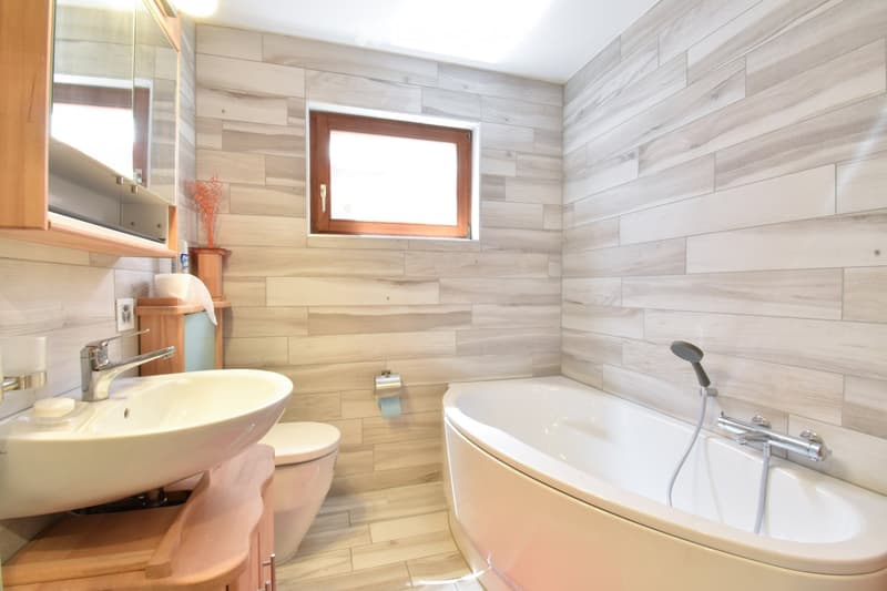 Modern bathroom with light wood paneling, a large window, a sink with a modern faucet, a toilet, a bathtub with a handheld showerhead, and a mirrored cabinet.