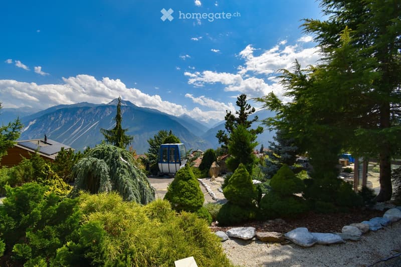 A well-maintained garden with neatly trimmed bushes and trees, featuring a cable car in the background and a scenic mountain view.