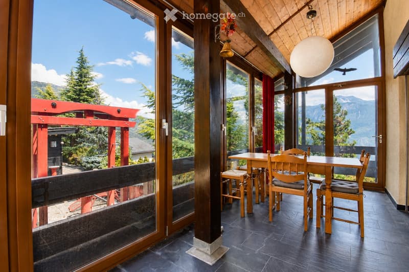 Bright, wooden-framed balcony with large windows offering mountain views, furnished with a wooden table and chairs.