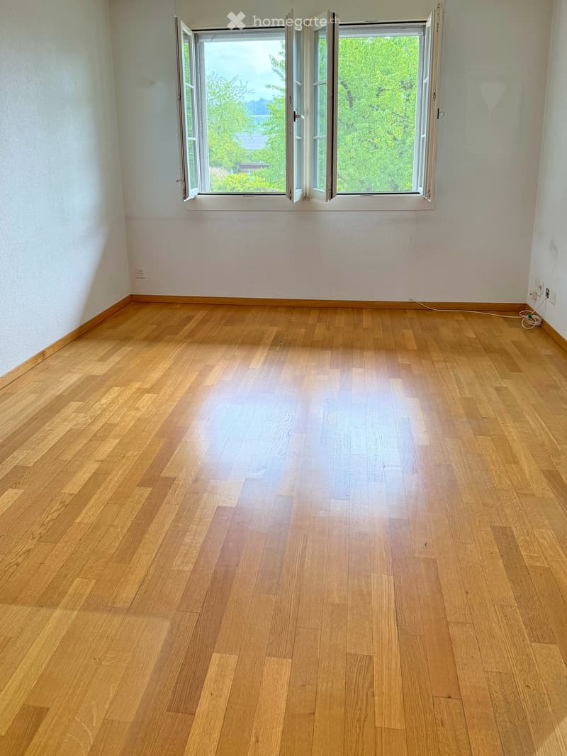 Empty room with light wooden flooring and white walls, featuring large windows with open shutters overlooking greenery.