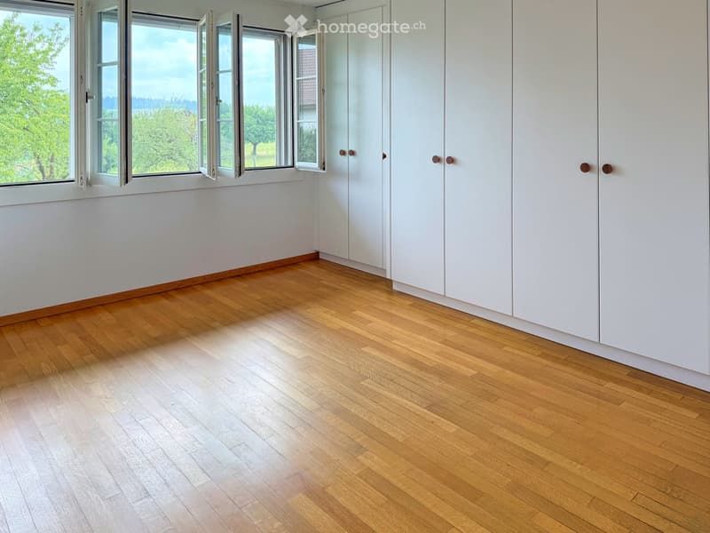 Empty room with light wooden flooring, white walls, and large windows with open shutters, revealing a green outdoor view.