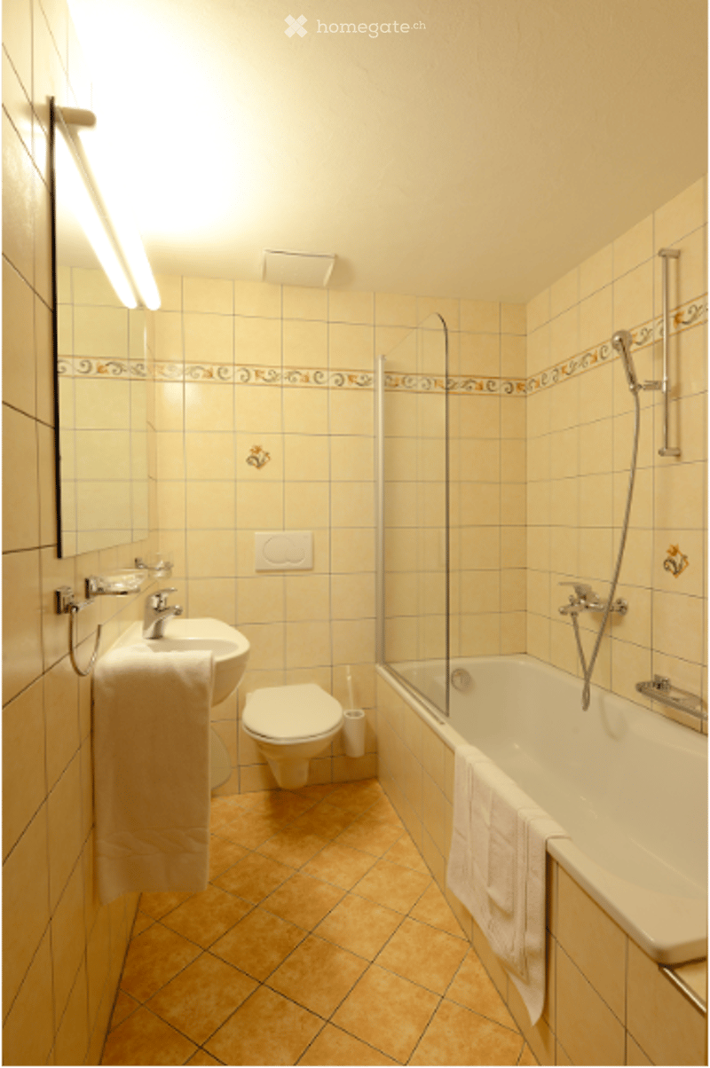 Bathroom with beige tiles, a sink with a mirror, a toilet, a bathtub with a glass shower enclosure, and a towel hanging on the side.
