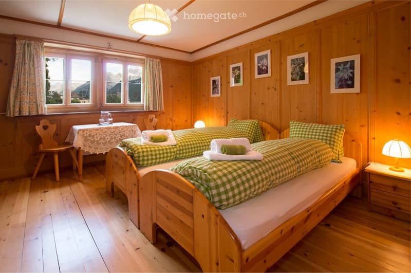 Cozy wooden bedroom with two single beds, nightstands, a small table, and a chair. The room features wooden walls, large windows with scenic views, and warm lighting.