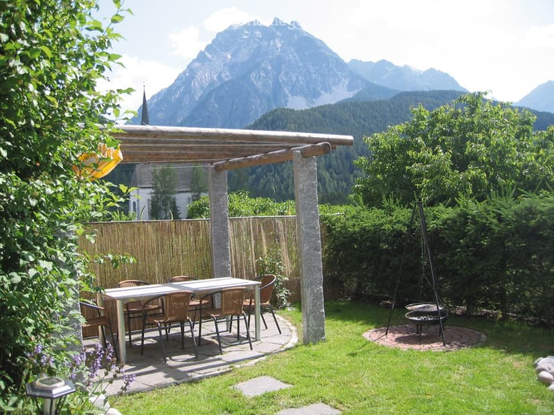 Outdoor patio with a table and chairs under a wooden pergola, surrounded by greenery and mountains.